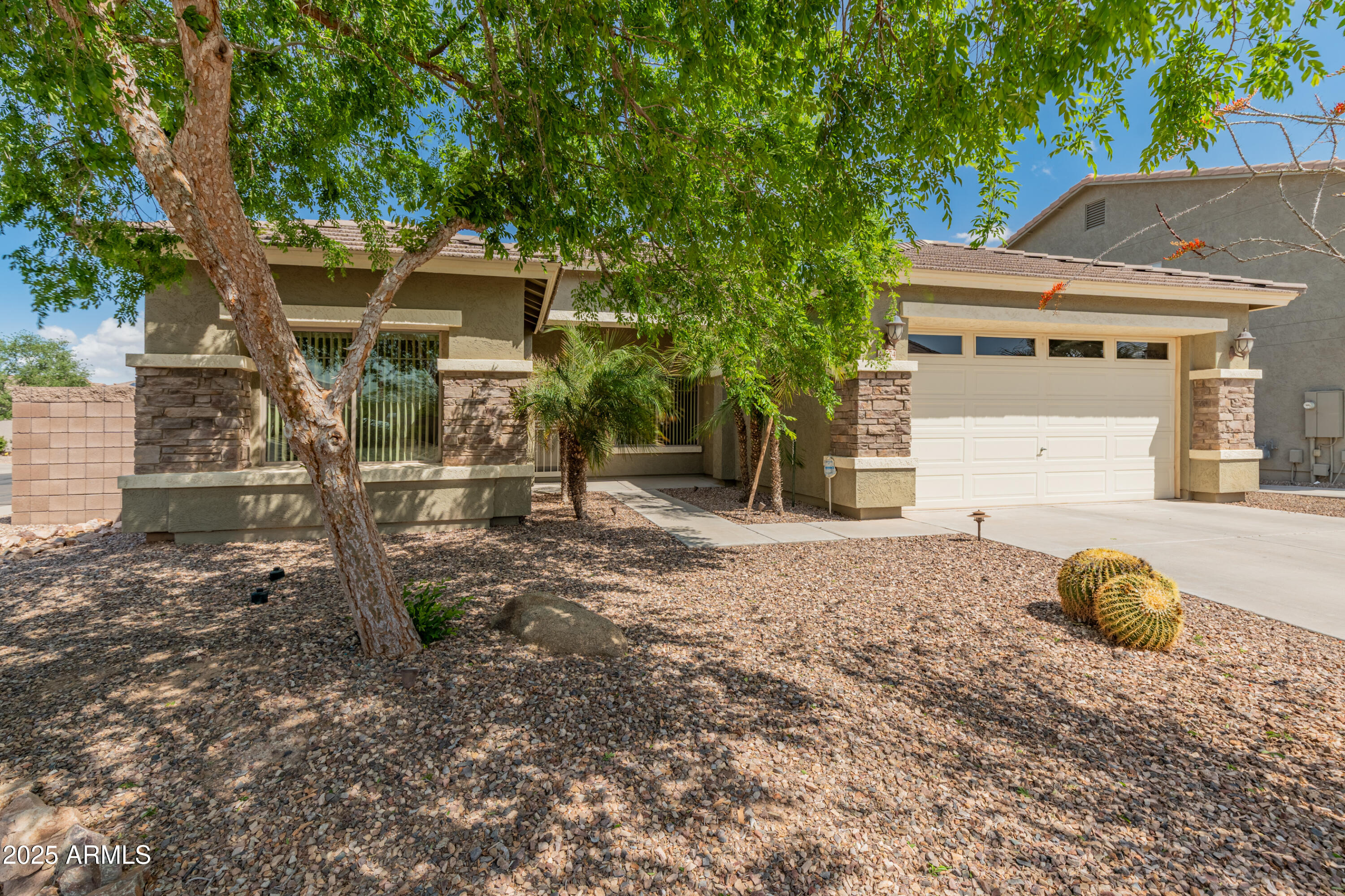 Single-story stucco home with stone accents, two-car garage, gravel yard, and a large shade tree.