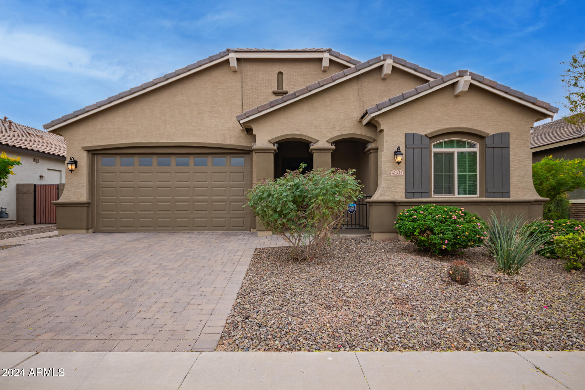 Tan stucco home with a two-car garage, covered entry, paved driveway, and desert landscaping.
