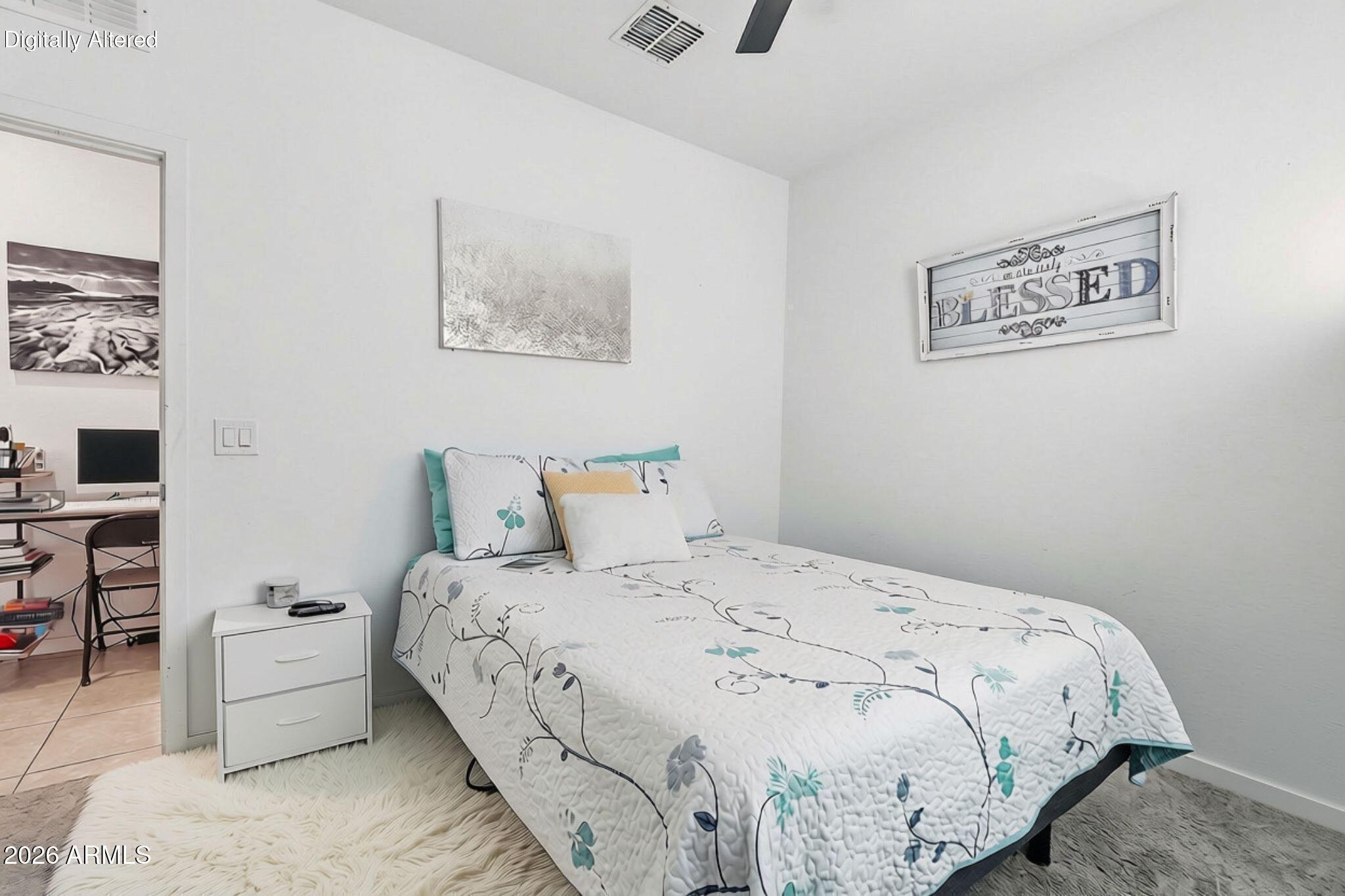 Bedroom with a white floral bedspread, turquoise accents, and a desk visible through the doorway.