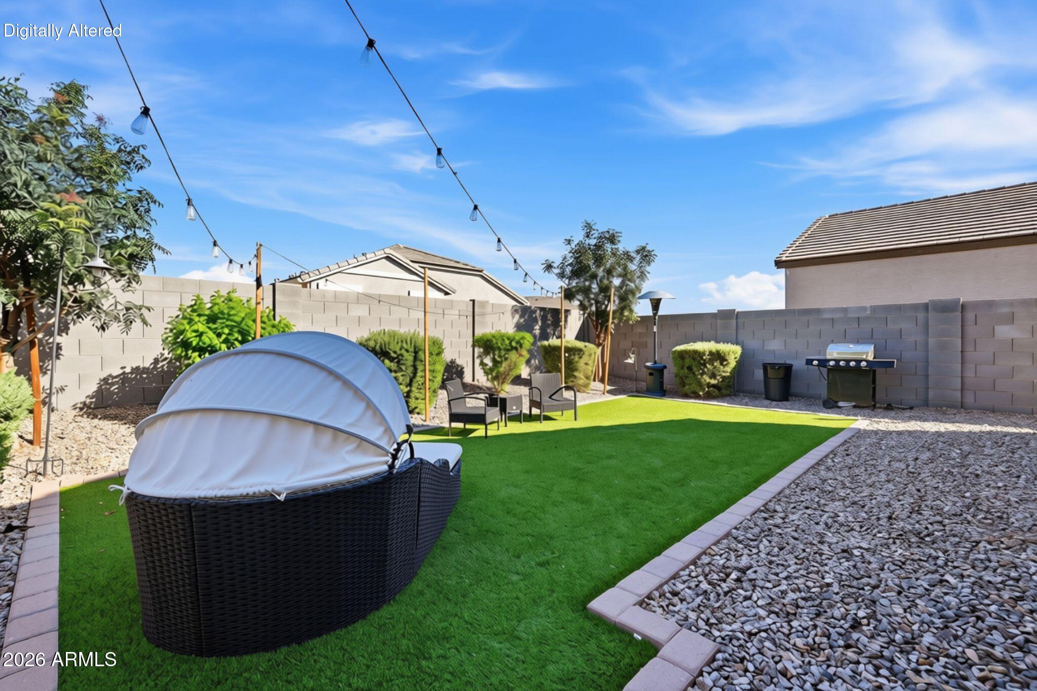 Backyard with artificial turf, covered wicker daybed, string lights, gravel beds, and barbecue.