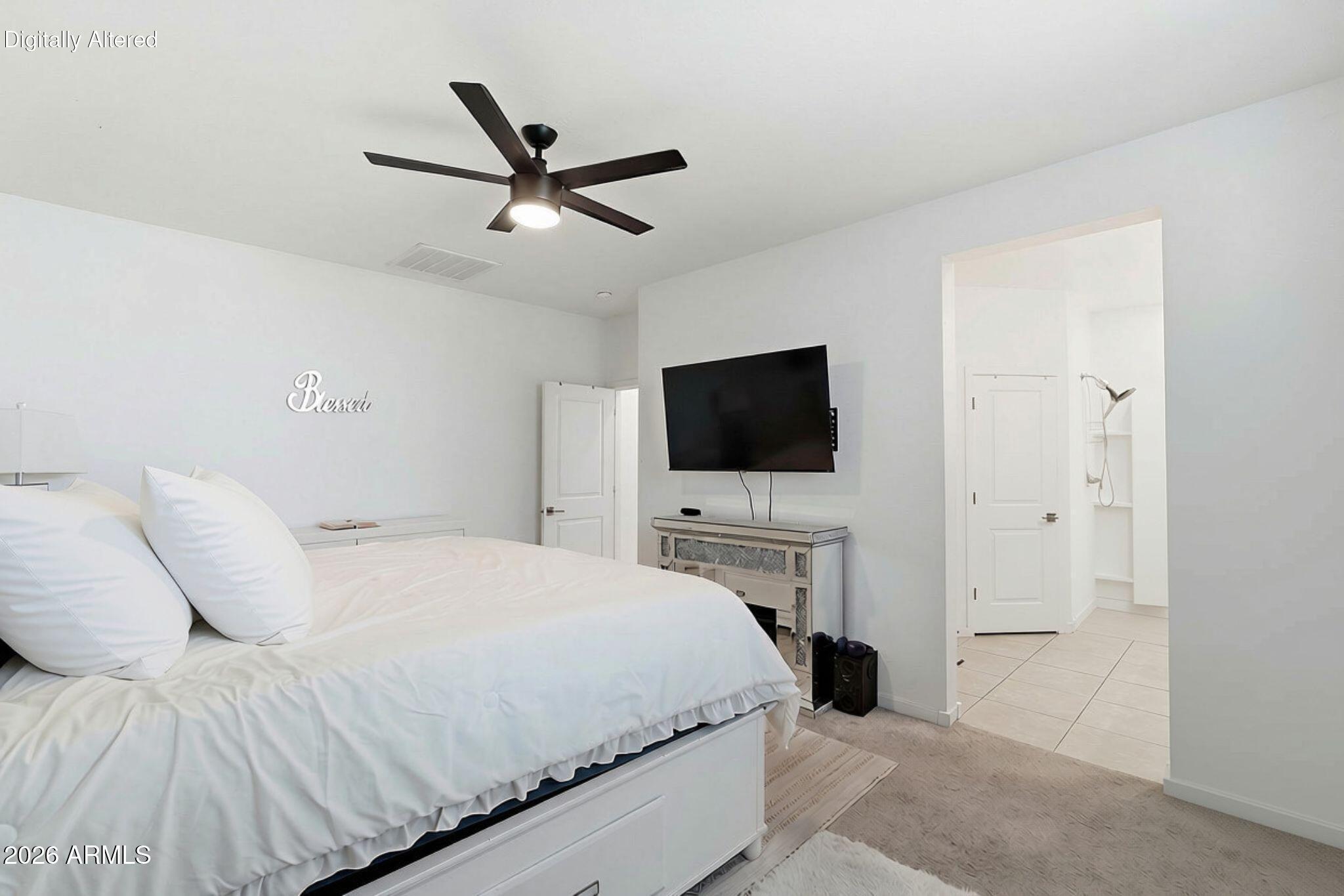 Bright bedroom with a white bed, dark ceiling fan, wall-mounted TV, and open tiled bathroom doorway.
