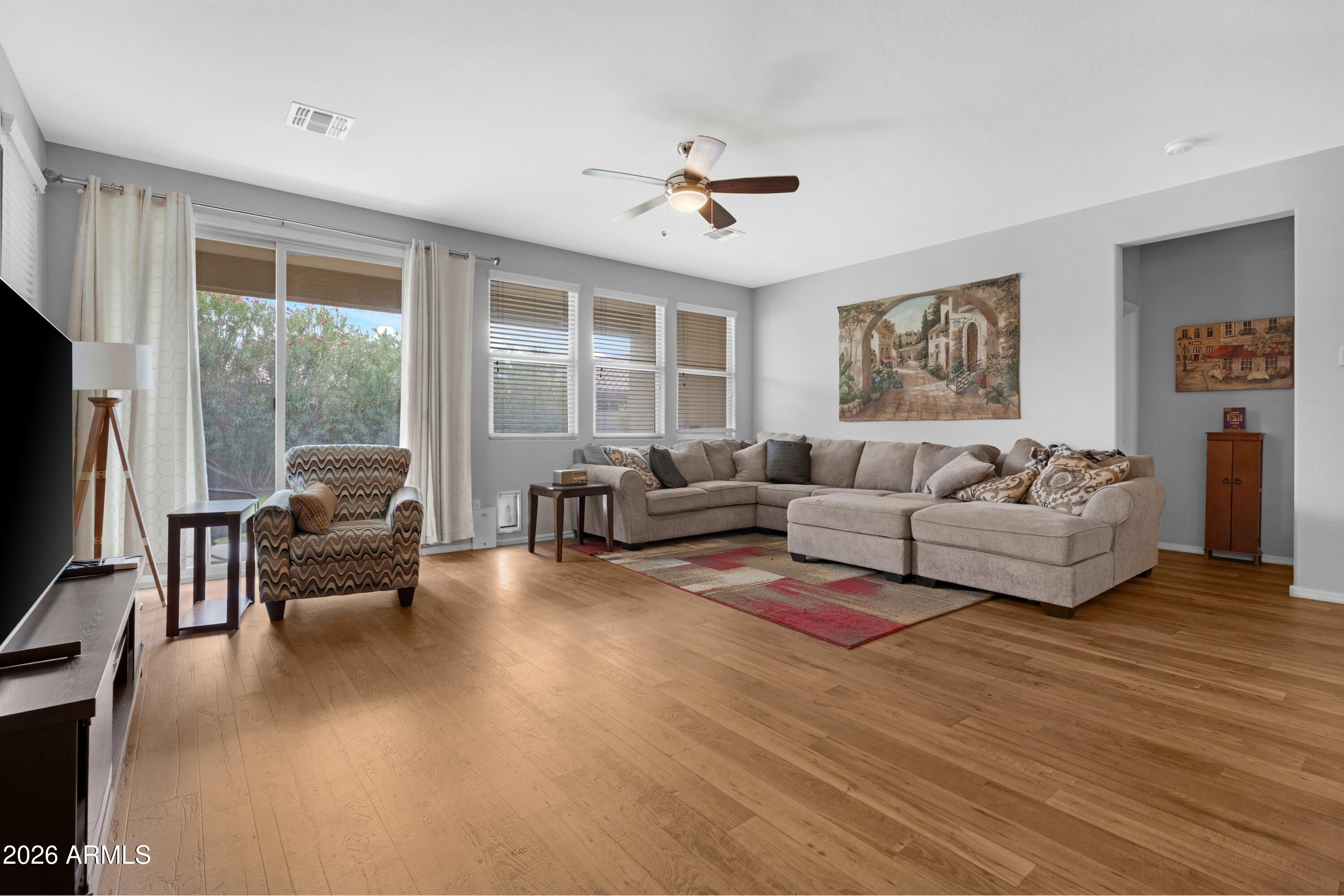 Living room with a beige sectional, patterned armchair, hardwood floors, and large curtained windows.