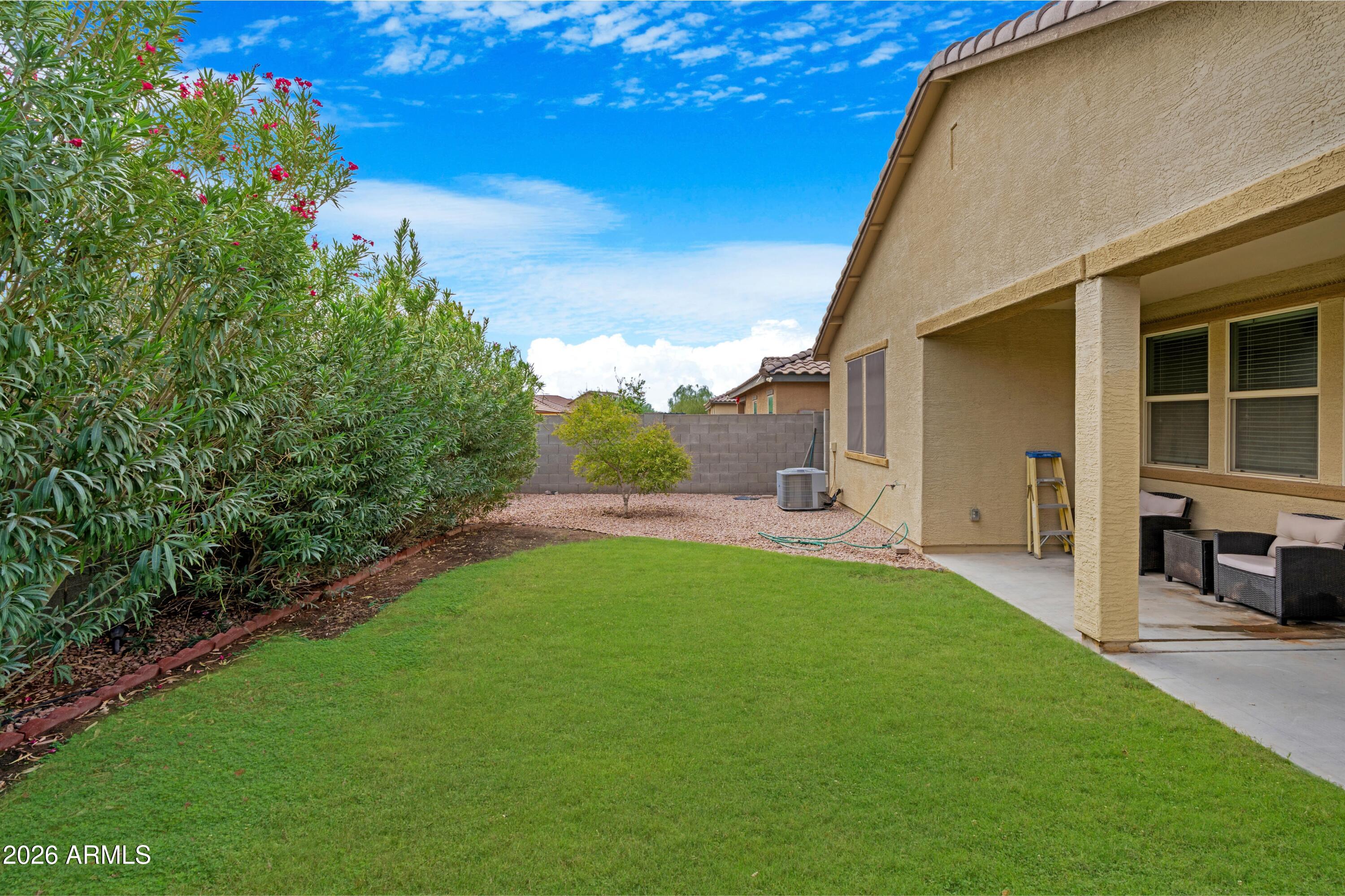 Backyard with a broad green lawn, flowering hedge, and covered patio with outdoor seating.
