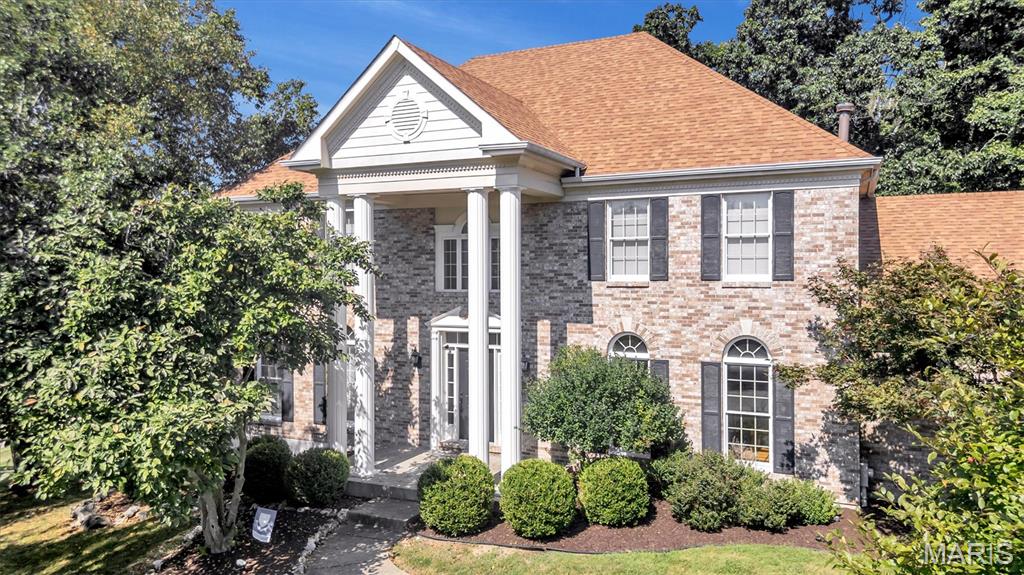 Two-story brick home with a white columned portico, tan roof, and landscaped front yard.