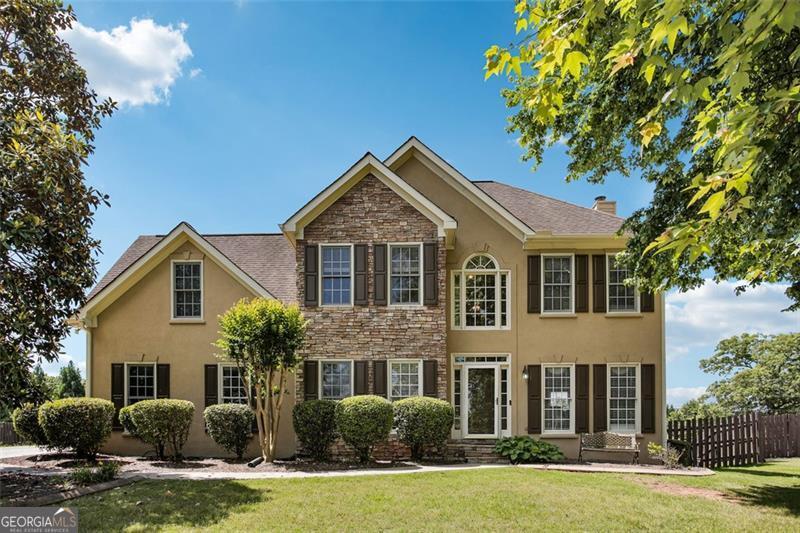 Two-story tan house with a stone-faced center, dark shutters, and a lawn framed by mature trees.