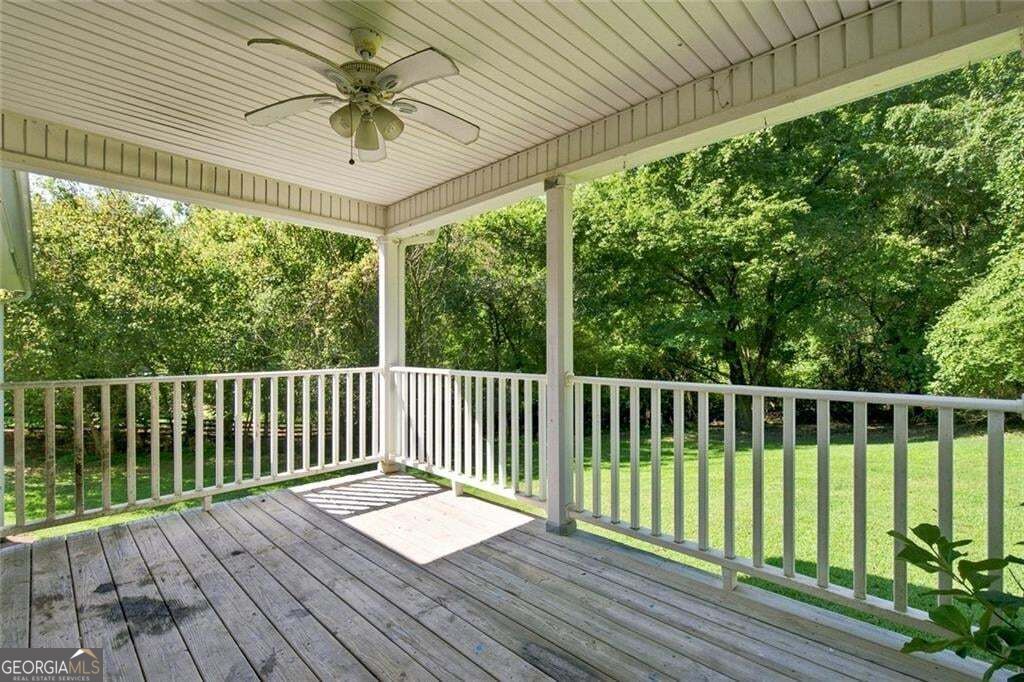 Covered wooden porch with a ceiling fan, white railing, and leafy yard beyond.
