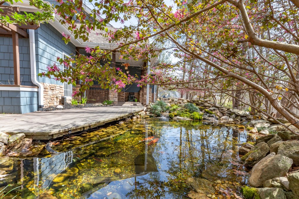 Blue-shingle home and wooden deck beside a clear, rock-lined pond beneath flowering branches.