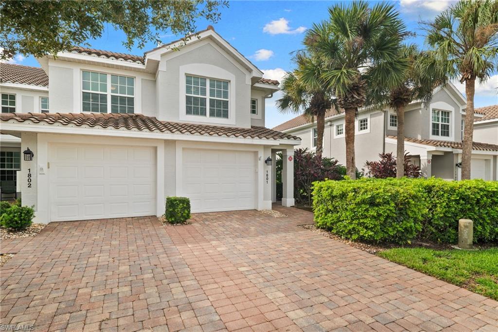 Two-story stucco home with twin garage doors, a brick-paver driveway, and palm landscaping.