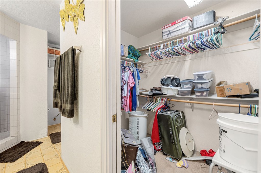 Walk-in closet with hanging clothes, storage bins, and luggage beside a tiled bathroom.