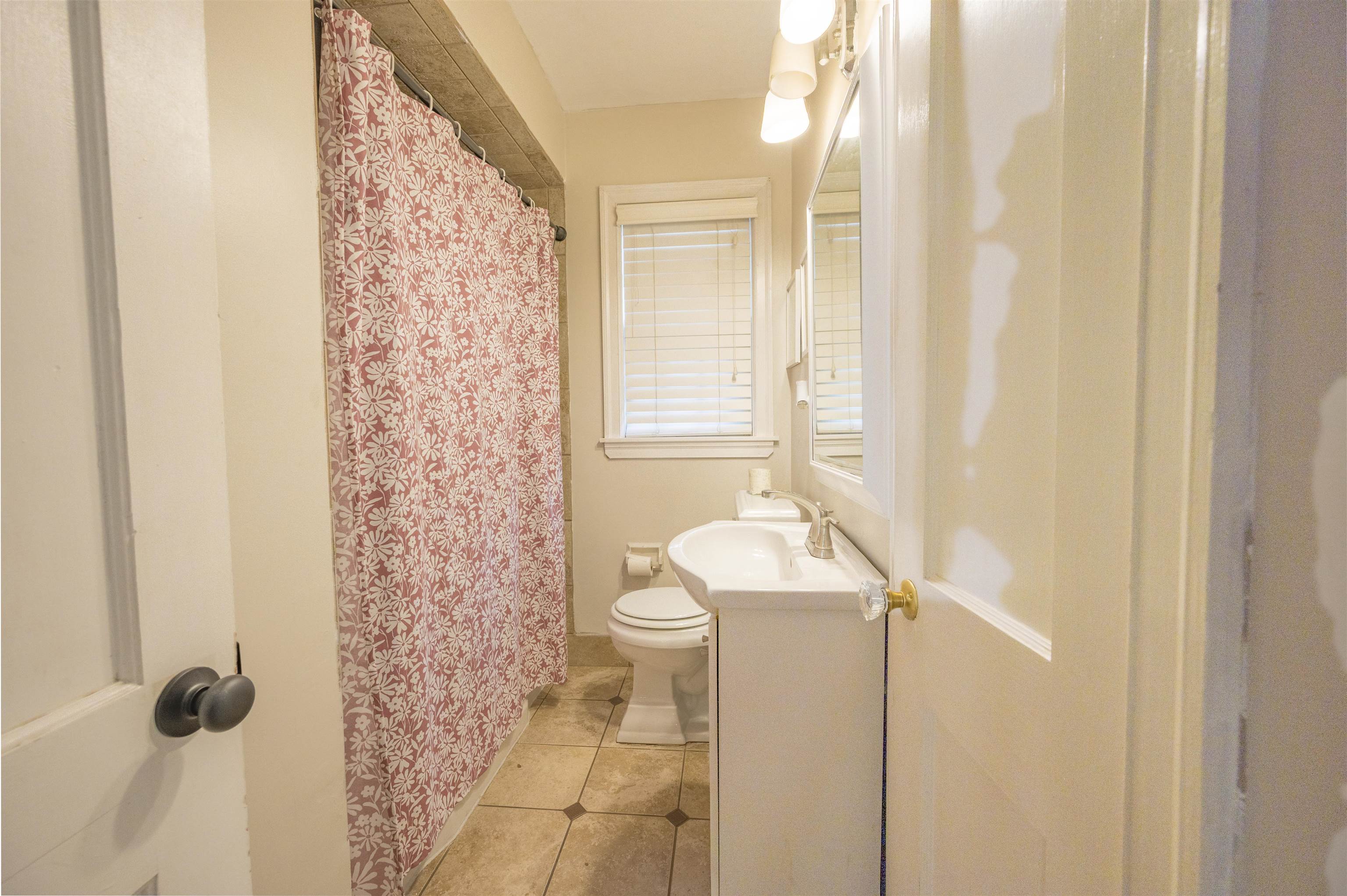 Bathroom with a pink floral shower curtain, tiled floor, and white vanity beneath a window.