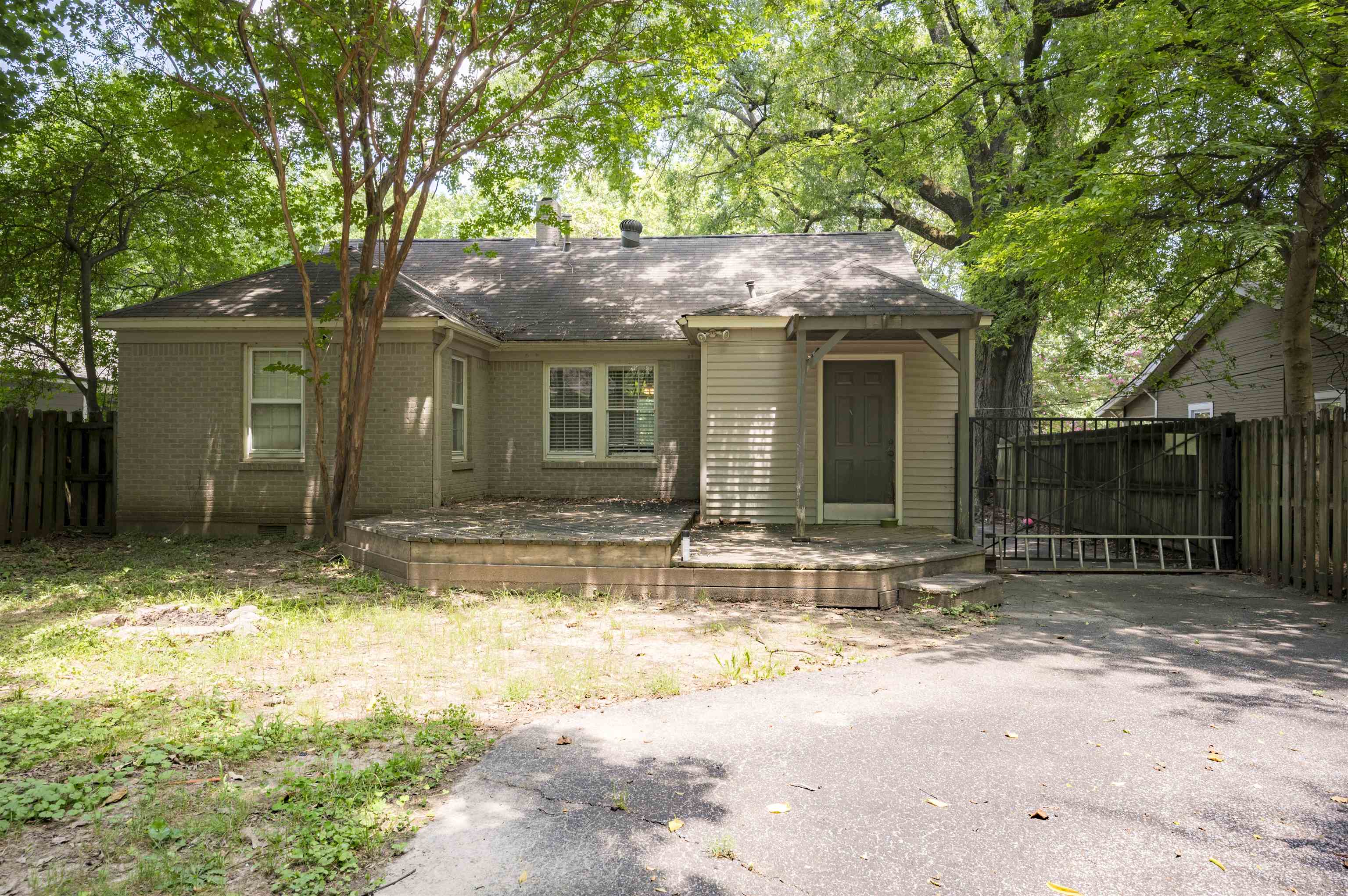 Rear exterior of a brick-and-siding house with a raised deck beneath mature trees.
