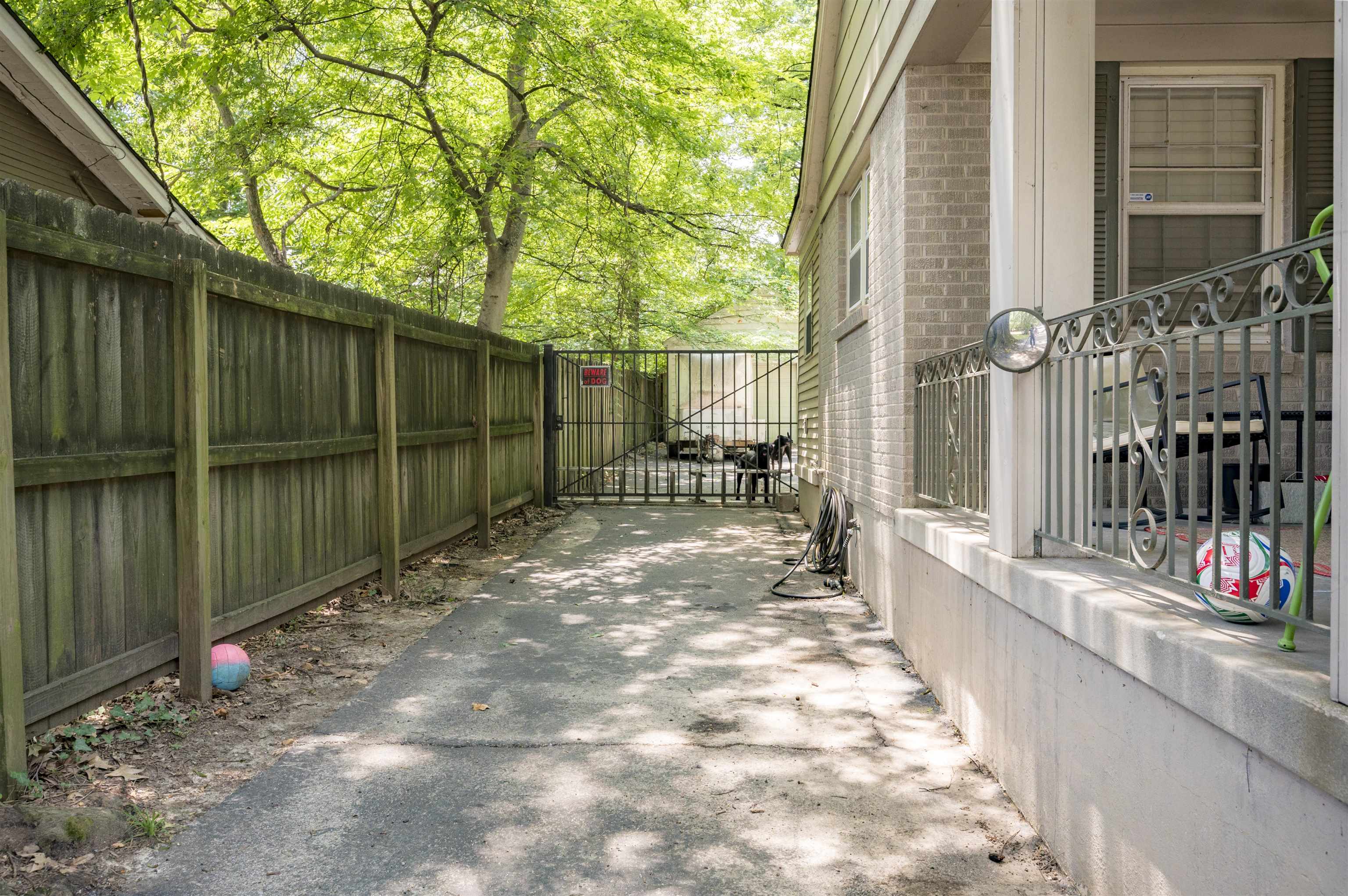 Concrete side walkway beside a brick house, bordered by a wooden fence and shaded by leafy trees.