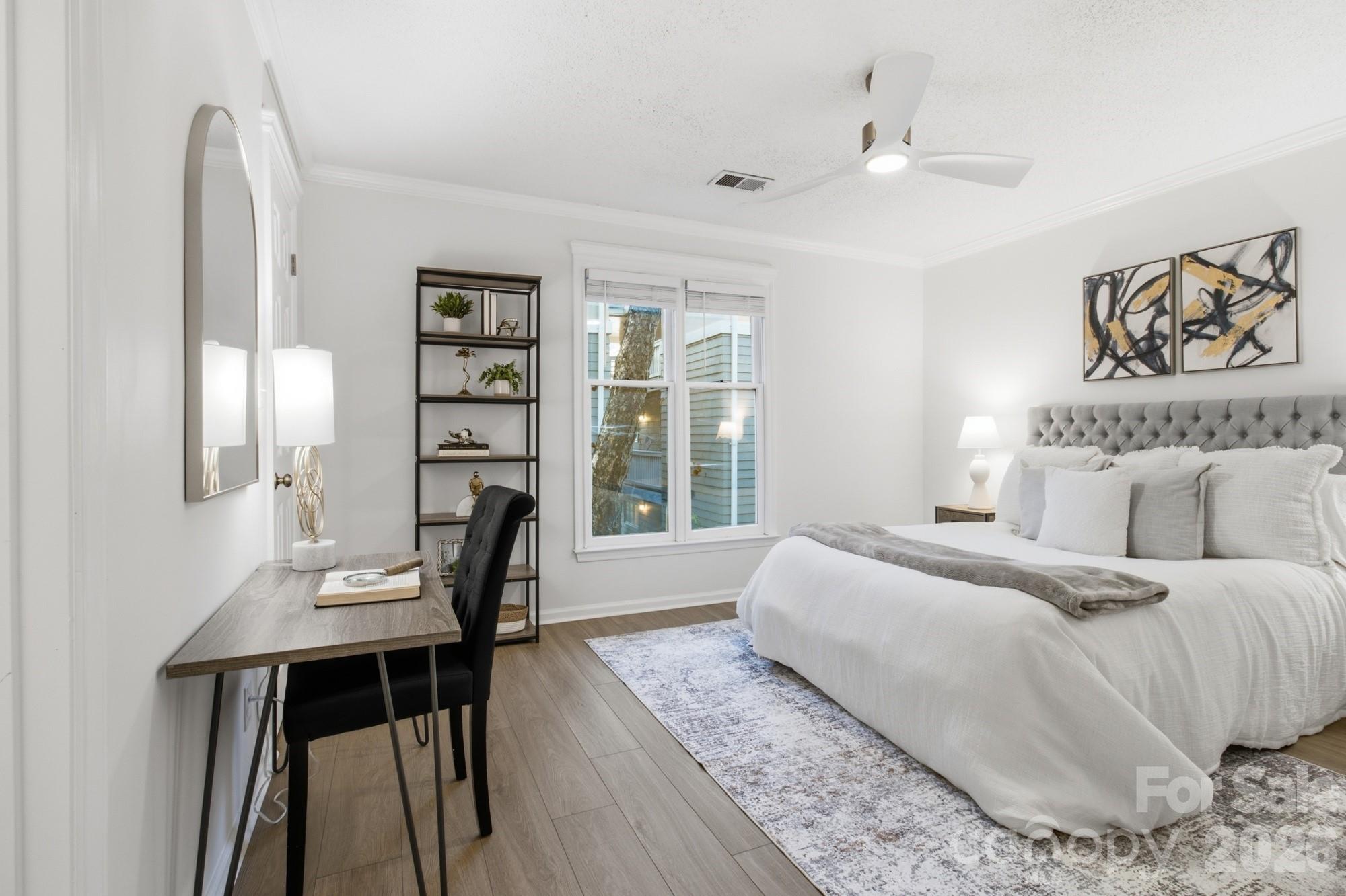 Staged bedroom with a white bed, tufted gray headboard, desk, shelving, and ceiling fan.