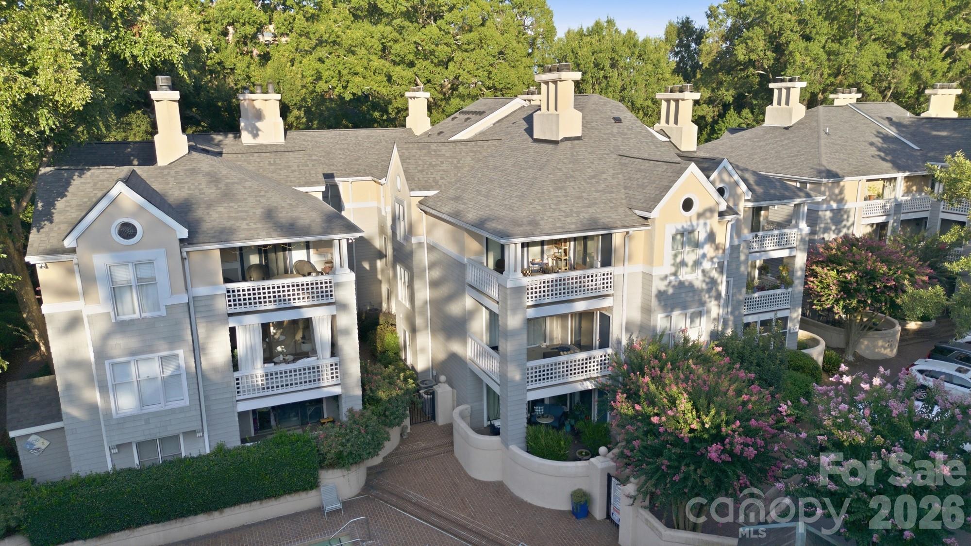 Condominium building with multiple balconies, surrounded by mature trees and pink flowering shrubs.