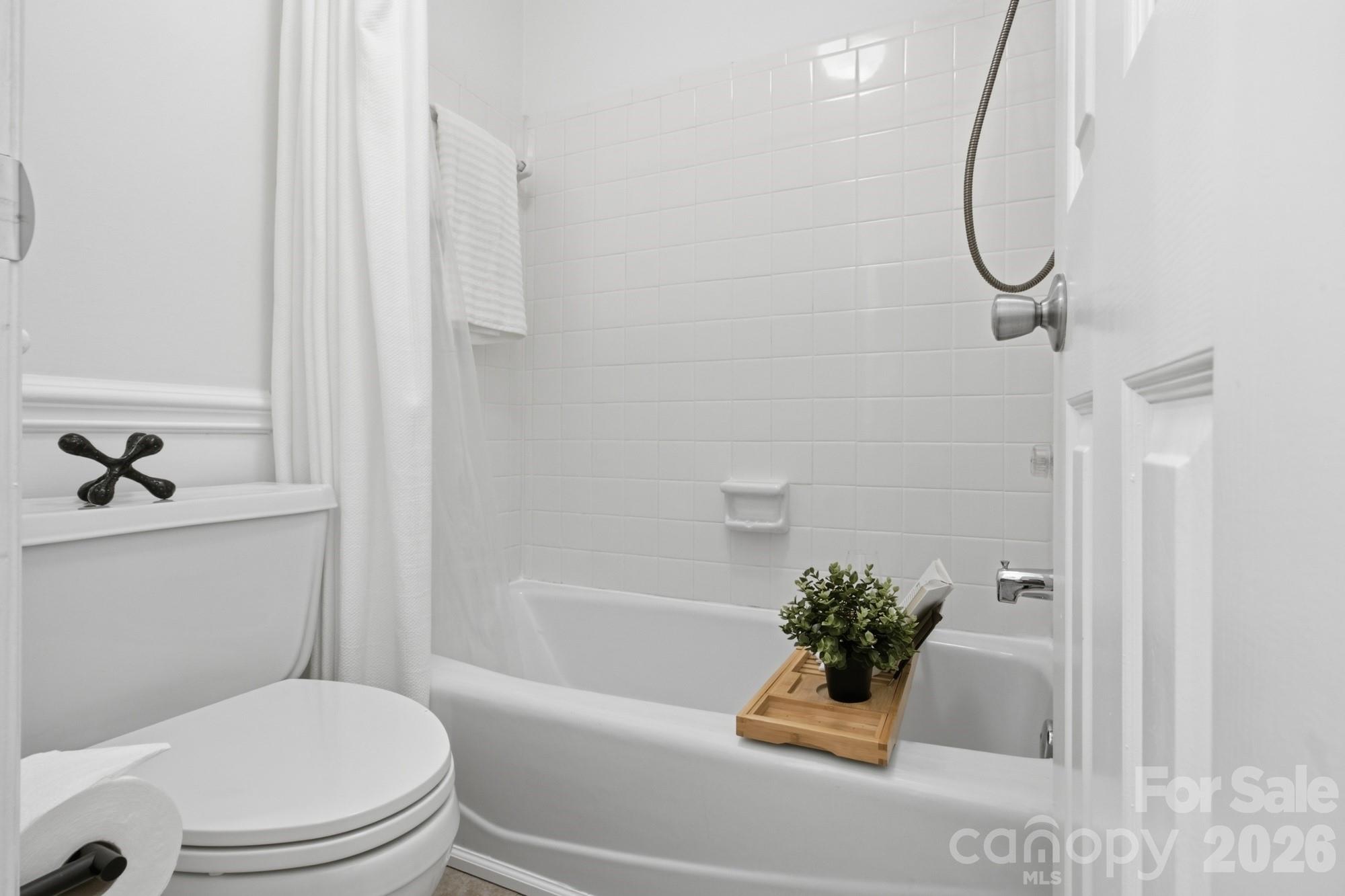 Bathroom with a white tiled tub and shower, toilet, and wooden bath tray with a potted plant.