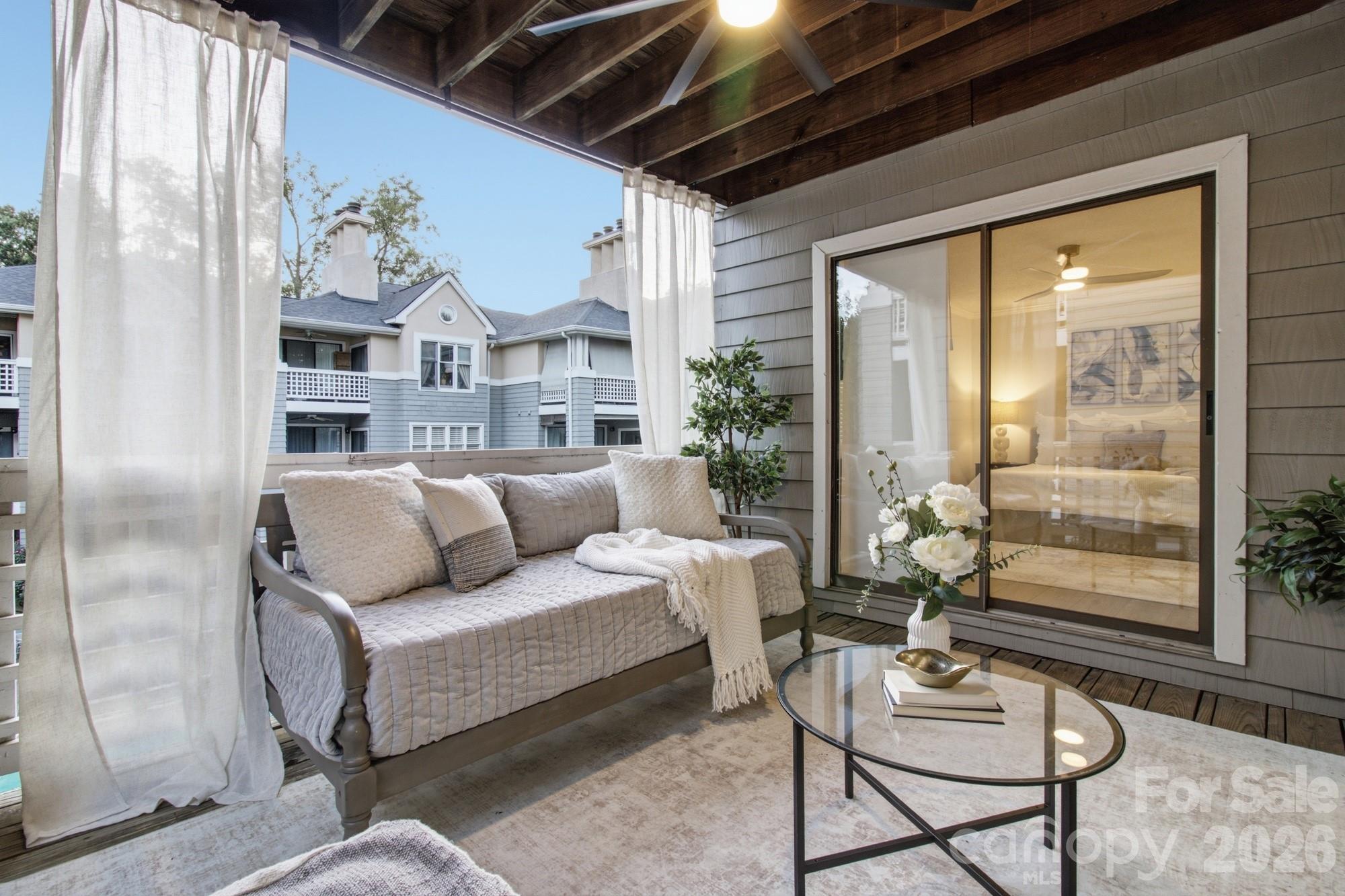 Furnished condo balcony with a cushioned sofa, sheer curtains, and views of neighboring buildings.