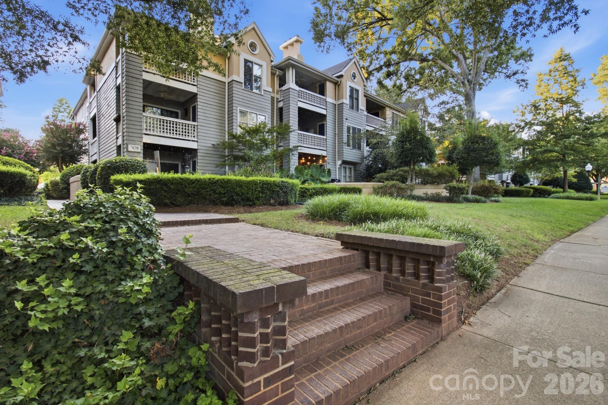 Multi-story condominium with gray siding and balconies, framed by landscaped grounds and brick entrance steps.