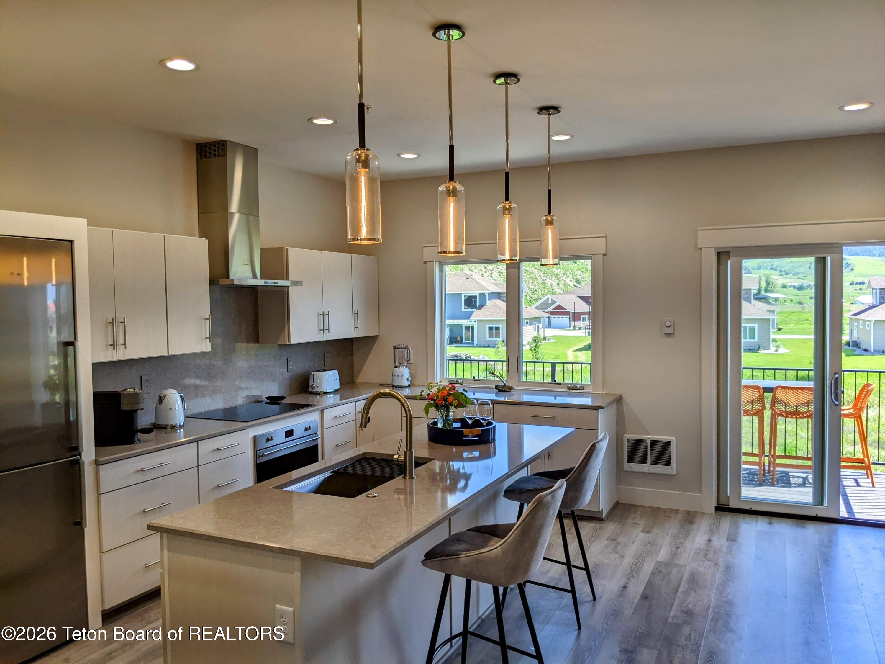 Modern kitchen with a large island and pendant lights, opening to a deck overlooking a sunny neighborhood.