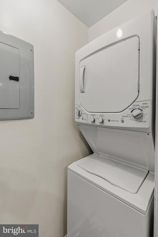 Stacked white washer and dryer in a narrow laundry closet beside a gray utility panel.