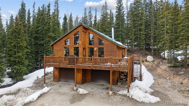 Log cabin with a raised deck and two-car garage among evergreen trees and patches of snow.