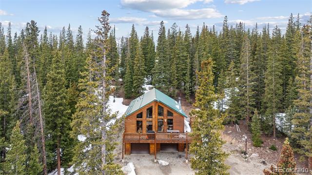 Wood cabin with a green roof and front balcony surrounded by snow-dusted evergreen forest.