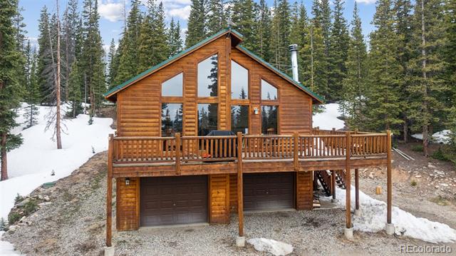 Log cabin with a large front deck and two garage doors among snowy evergreens.