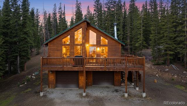 Log cabin with a raised front deck and two garage doors among evergreen trees at dusk.