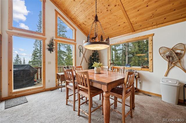 Dining area with a large wooden table, vaulted pine ceiling, and tall windows overlooking trees.