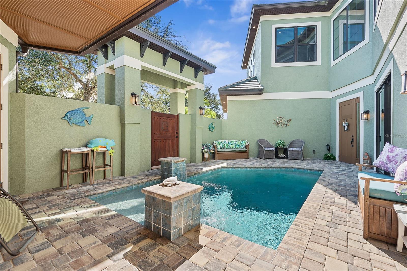 Pool courtyard with a paver deck, covered patio, and green two-story home.