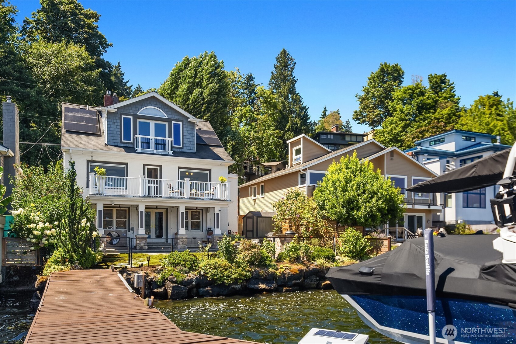 Waterfront home with a wooden dock, landscaped shore, and neighboring houses.