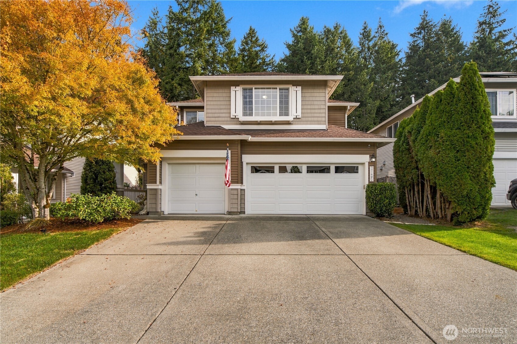 Two-story beige house with double garage, broad driveway, American flag, and golden autumn tree.