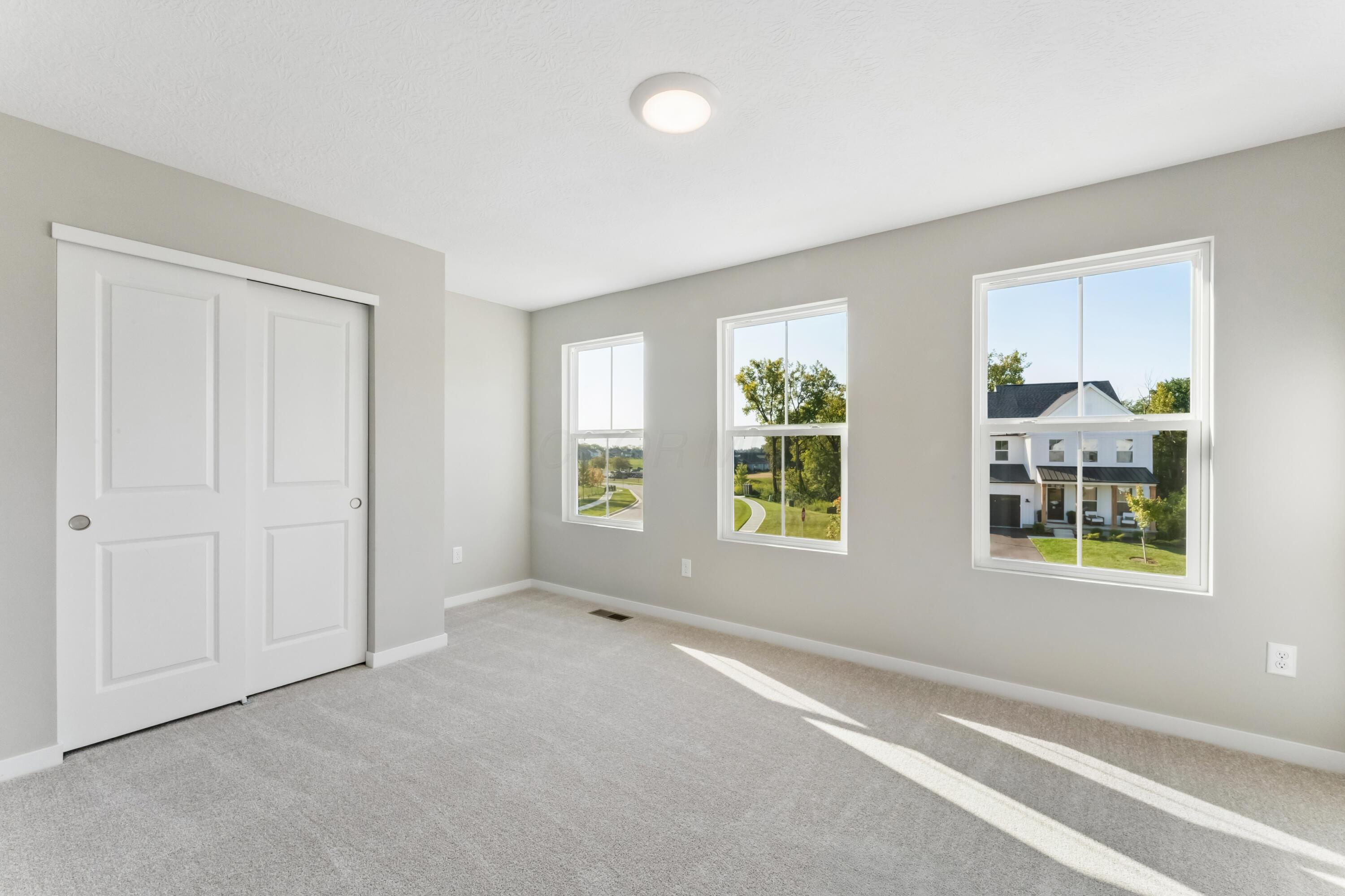 Empty bedroom with a sliding-door closet, three windows, and sunlight across the carpet.