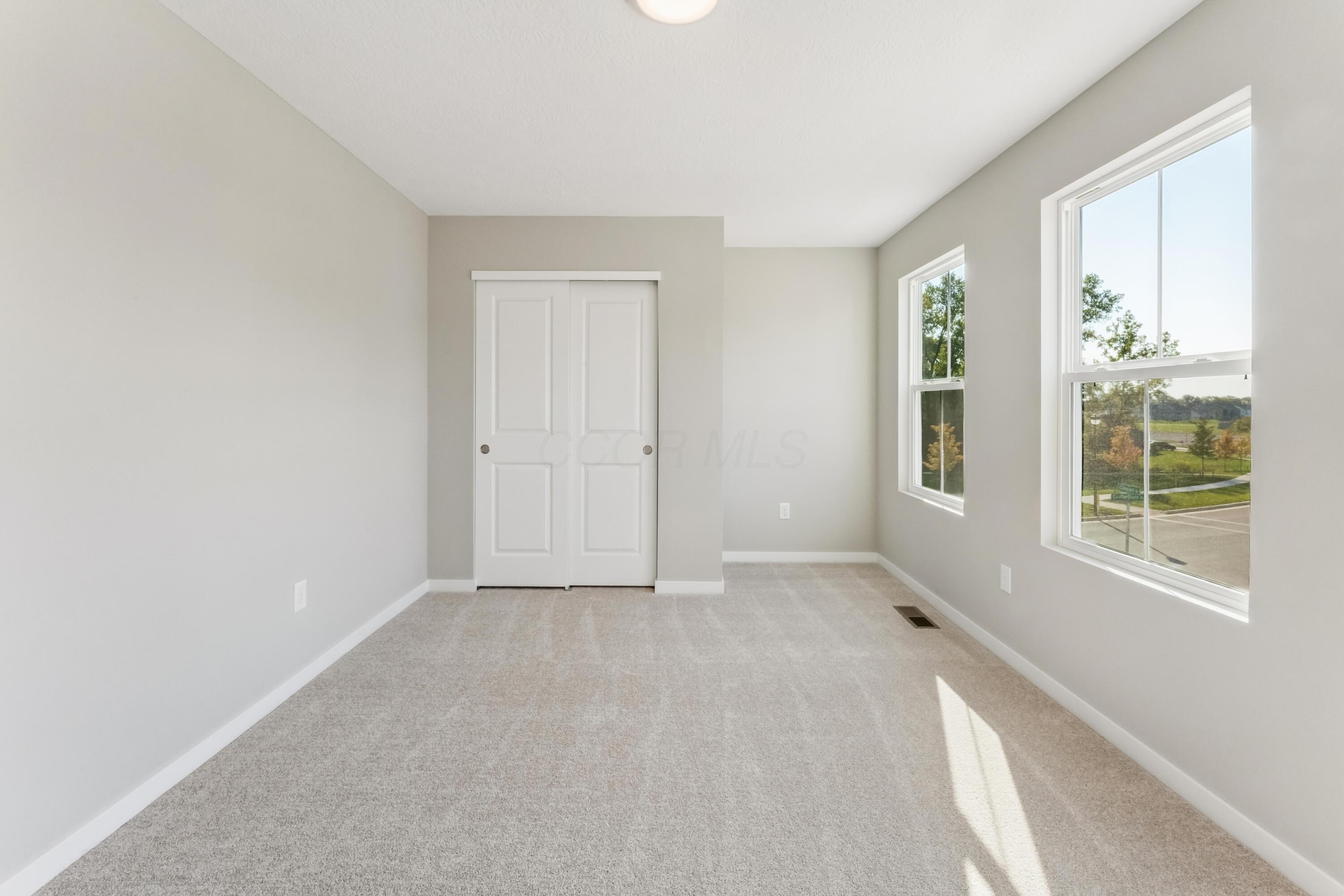 Vacant carpeted bedroom with a double-door closet and two large sunlit windows.