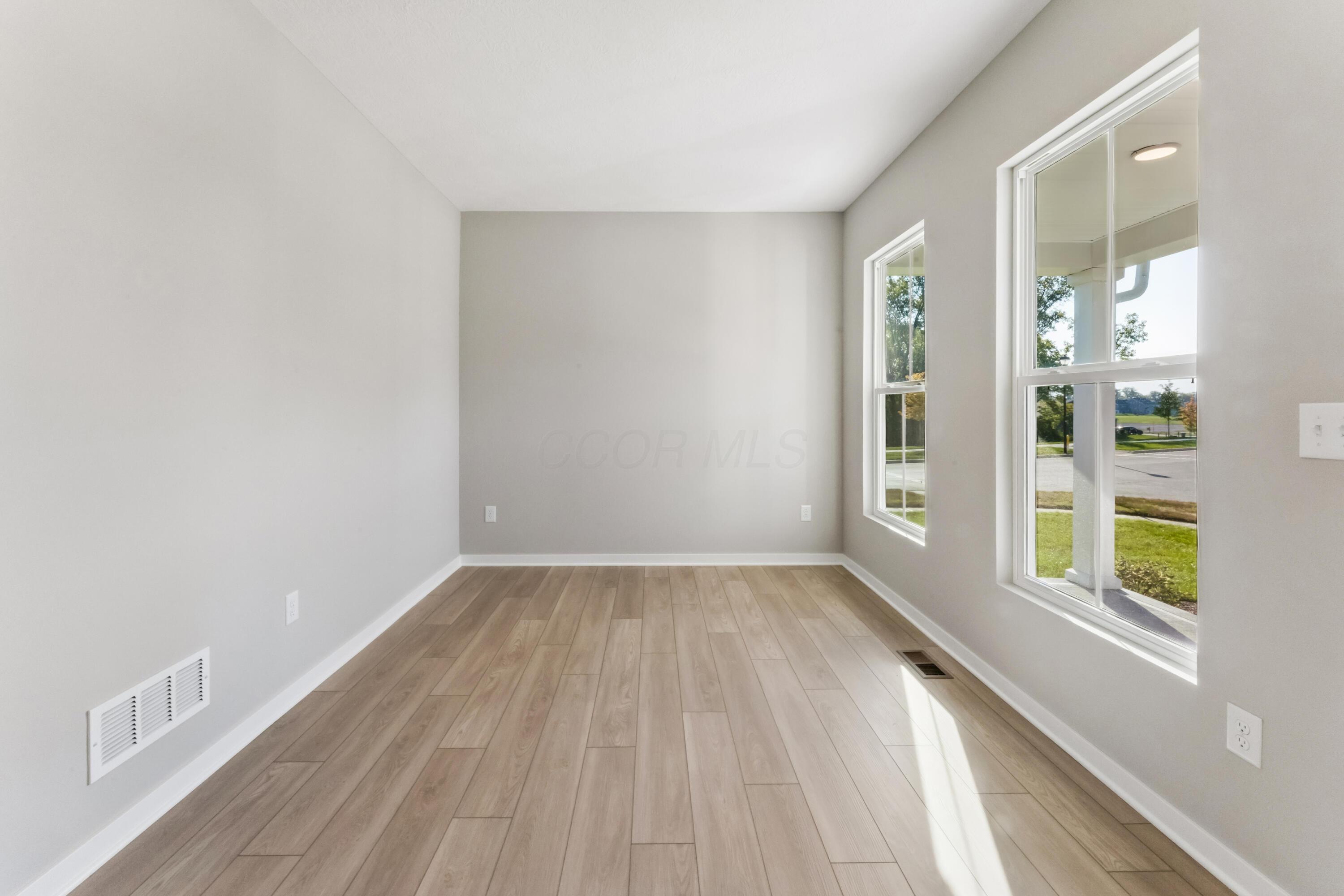 Empty room with wood-look plank flooring and large windows overlooking a lawn and road.