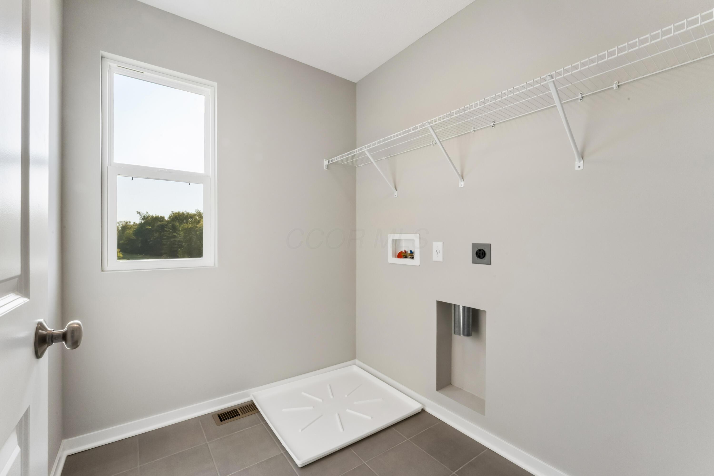 Empty laundry room with gray tile, high window, wire shelving, washer hookups, and an appliance pan.