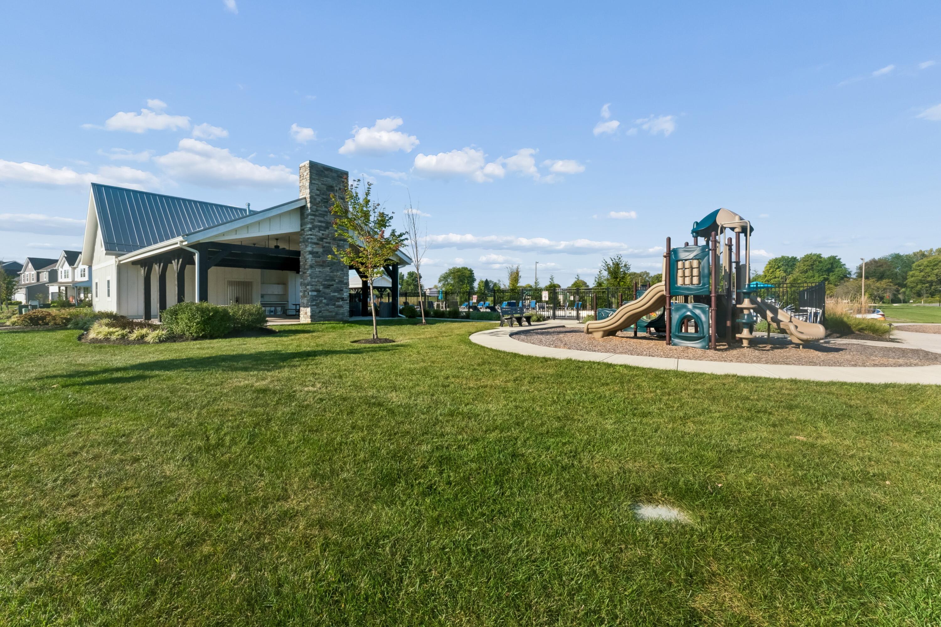 Community pavilion and fenced playground with slides across a broad green lawn.