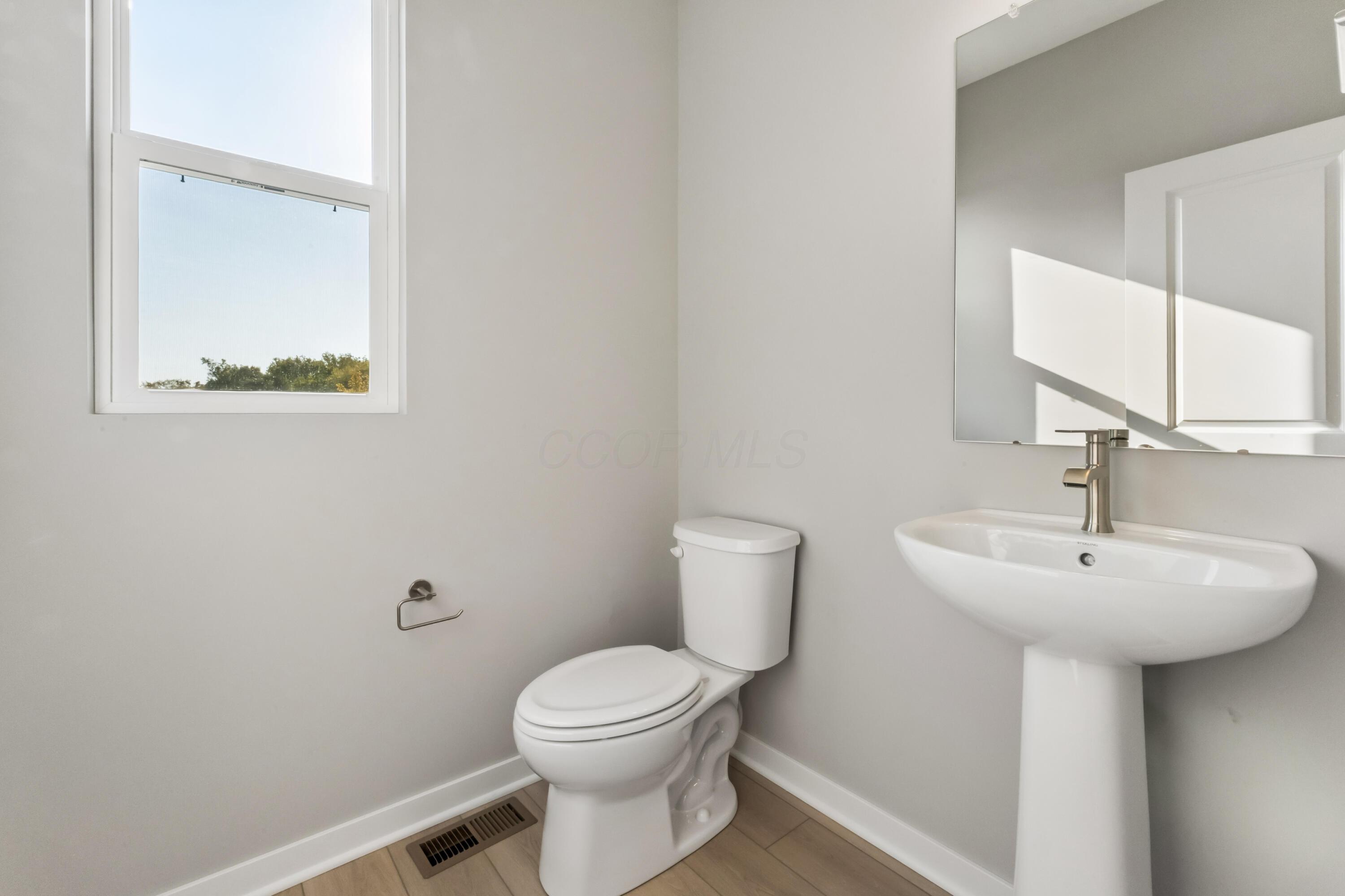 Bathroom with a pedestal sink and toilet beneath a large mirror, lit by daylight from a window.