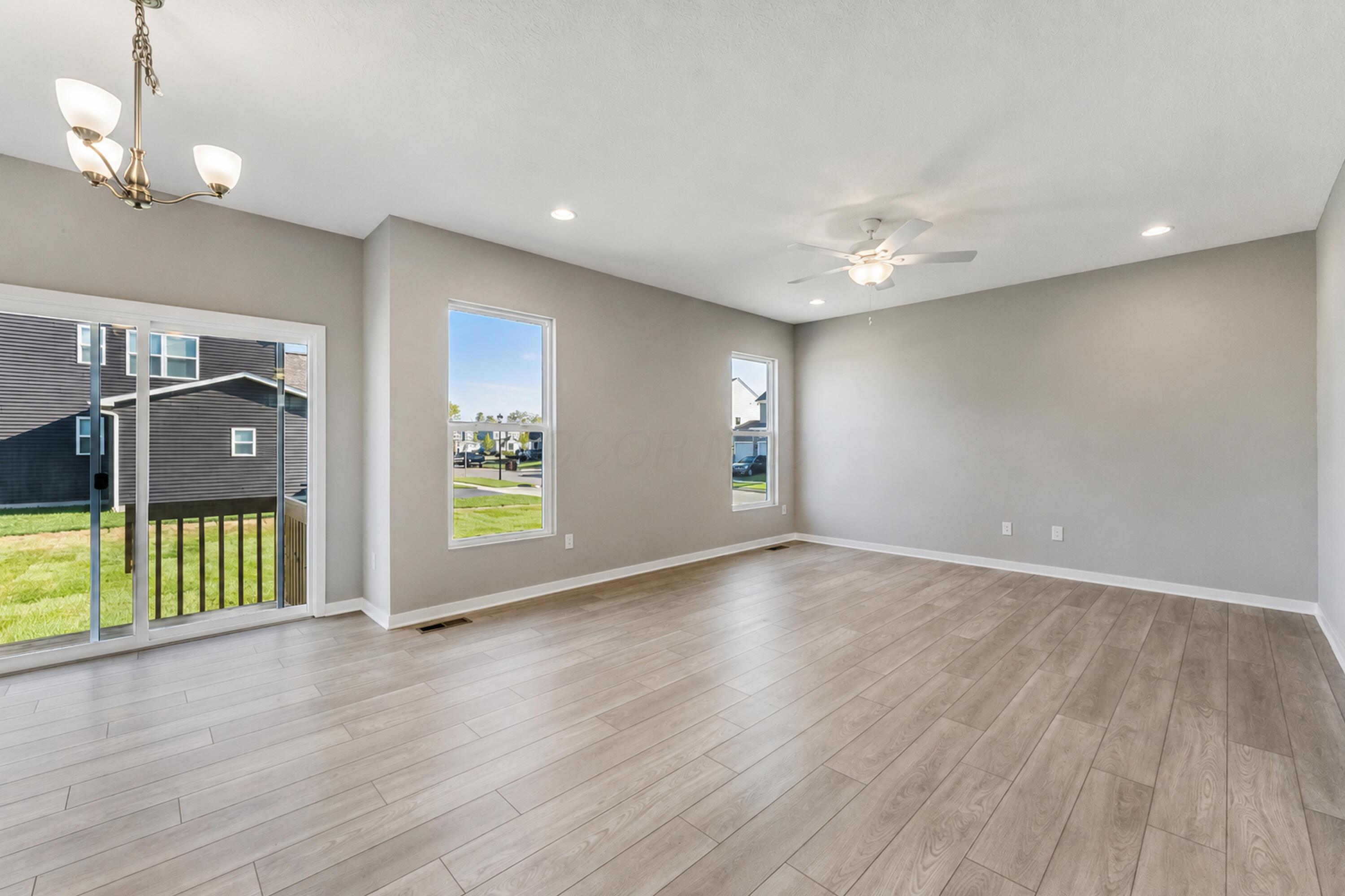 Empty living room with pale plank flooring, sliding glass doors, windows, and a ceiling fan.
