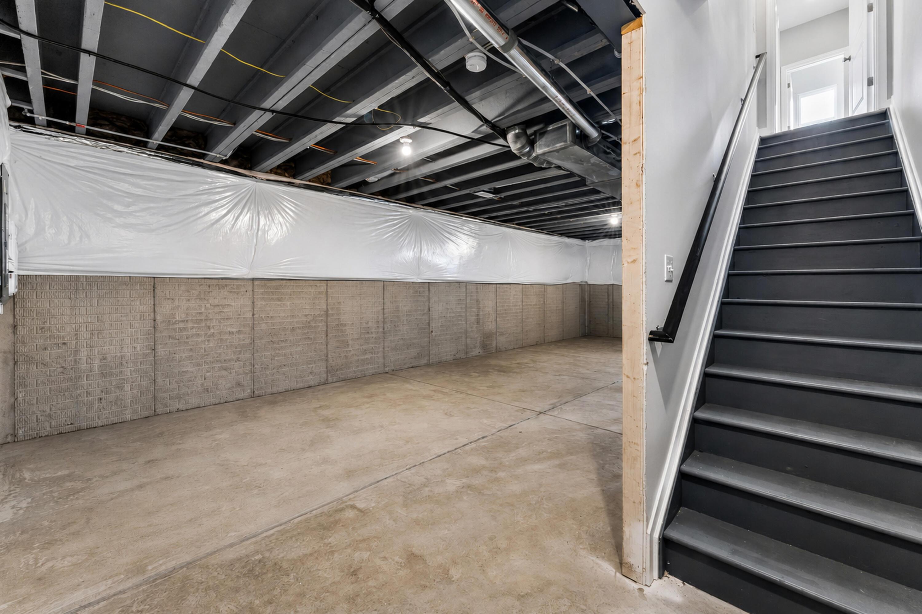 Unfinished basement with exposed ceiling joists, plastic-covered walls, and a staircase.