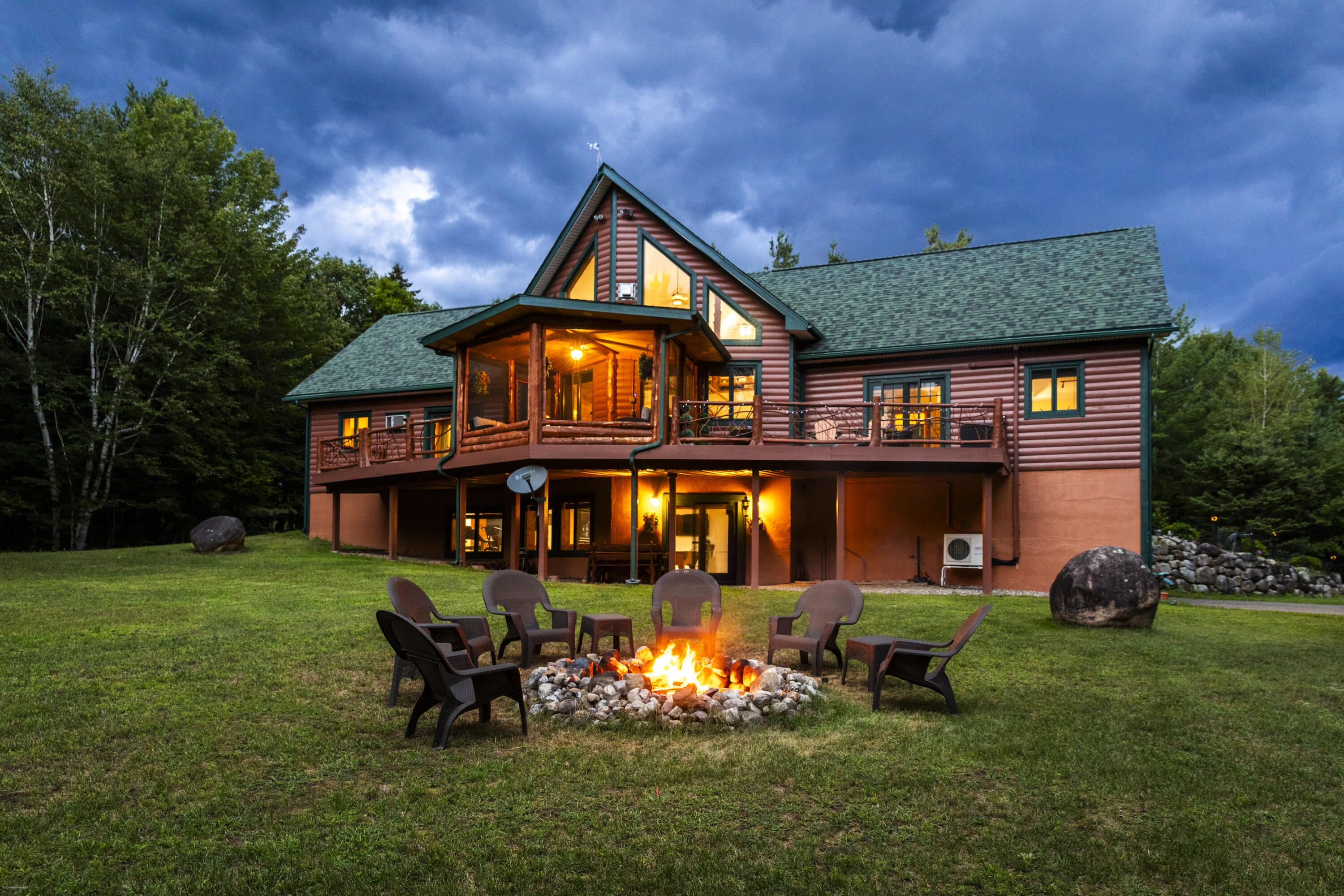 Log-sided home with a wraparound deck and glowing windows, behind a lawn fire pit and Adirondack chairs.