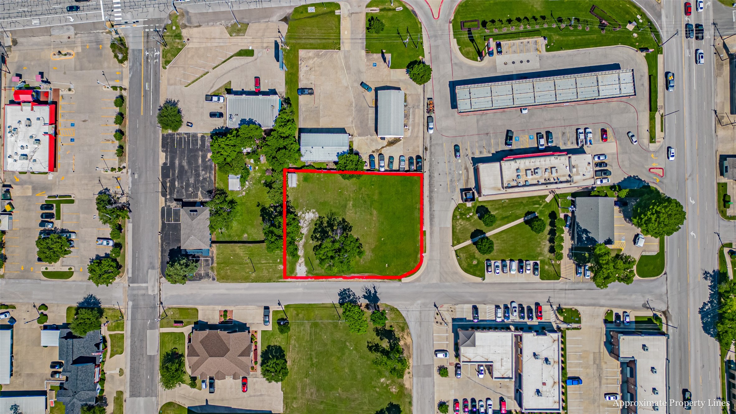Aerial view of a grassy vacant lot outlined in red among surrounding businesses and parking lots.