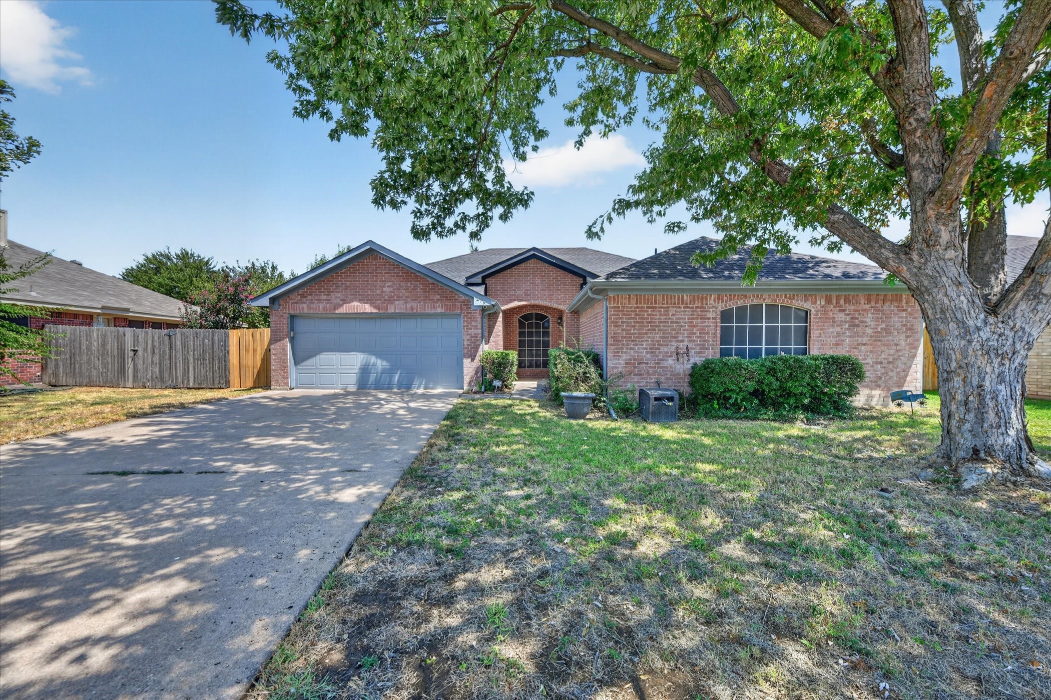 Red brick home with a gray garage and arched entry beneath a large shade tree.
