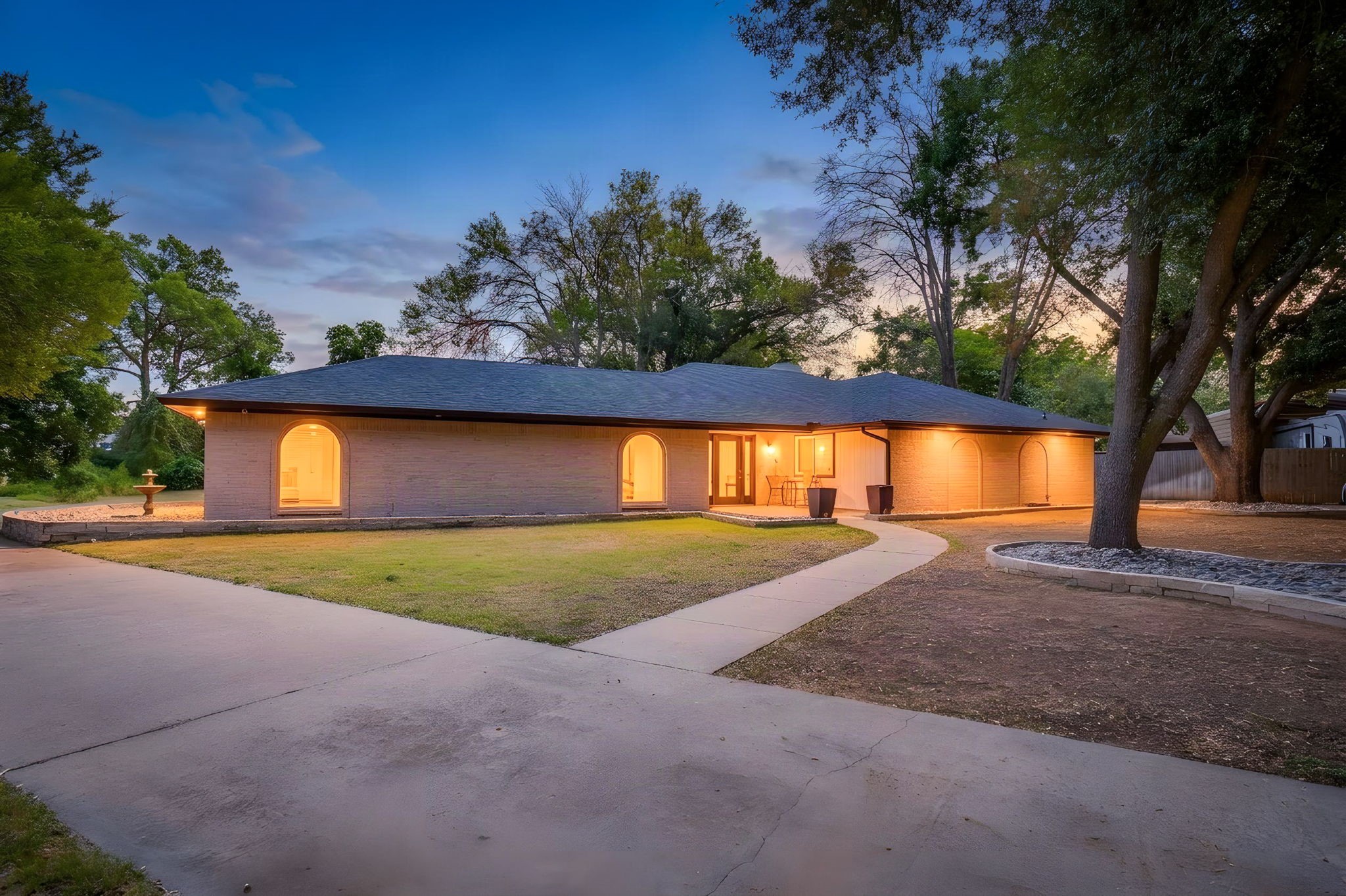 Single-story brick house at dusk with arched windows, warm lights, and a curved walkway.