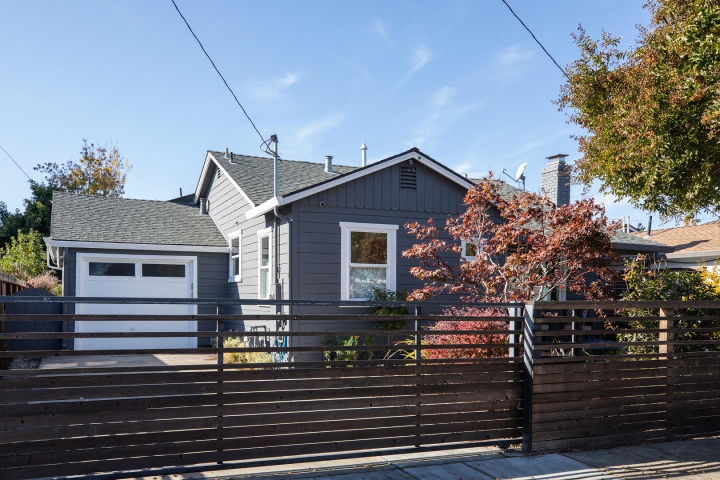Gray house with an attached garage behind a dark wooden fence, framed by red autumn foliage.