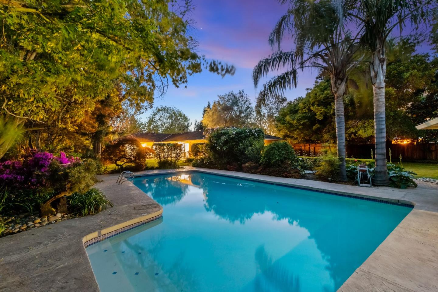 Backyard swimming pool at dusk, framed by lush landscaping and palm trees.