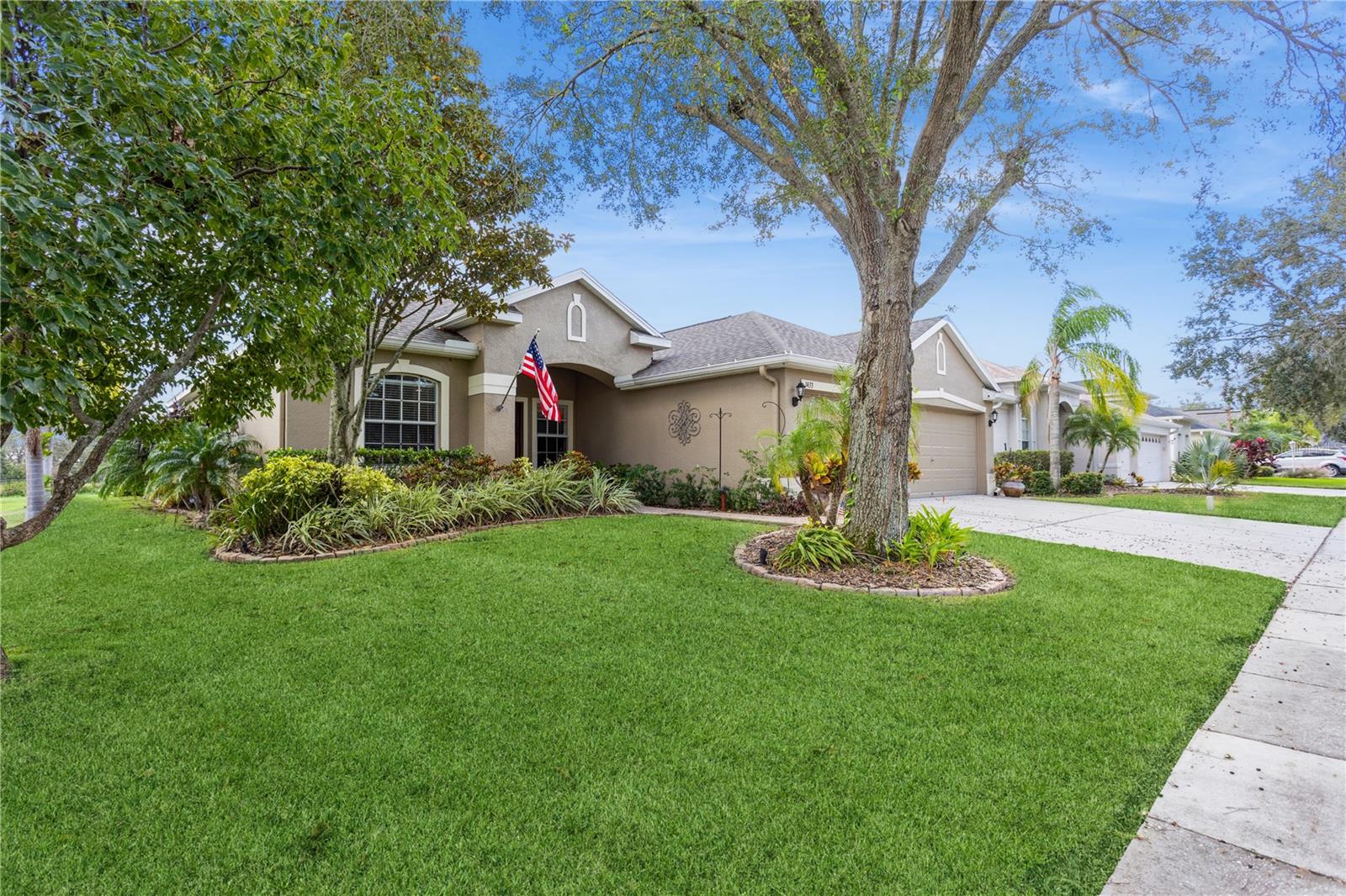 Single-story stucco home with an American flag, landscaped front yard, and mature shade trees.