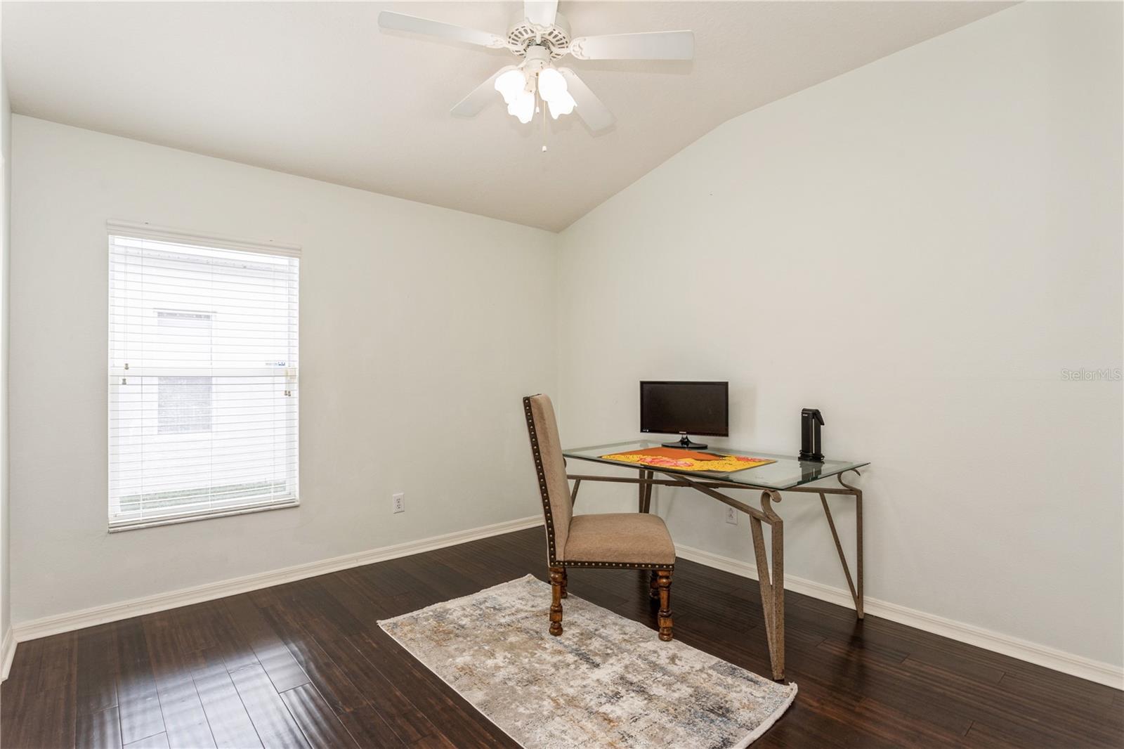 Home office with a glass-top desk, upholstered chair, dark wood floors, and a ceiling fan.