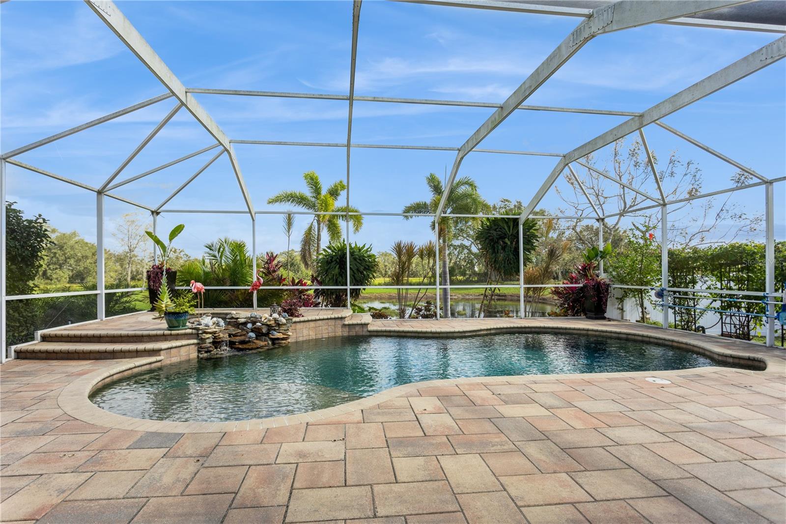 Screened swimming pool with a curved paver deck, rock waterfall, and tropical plants beside a pond.