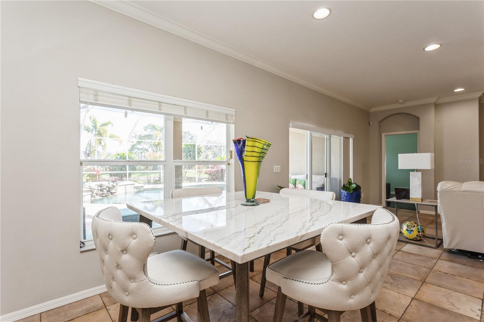 Dining area with a marble-look table and upholstered chairs overlooking a backyard pool.