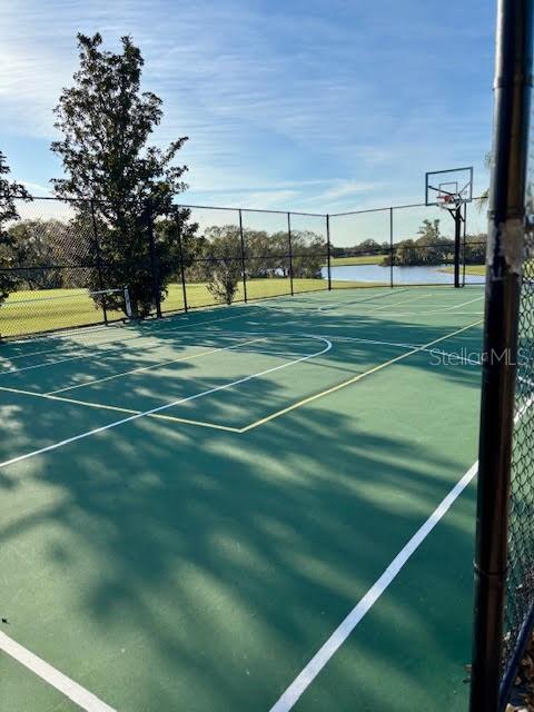 Fenced outdoor basketball court beside a pond, with a hoop and tree shadows across the playing surface.